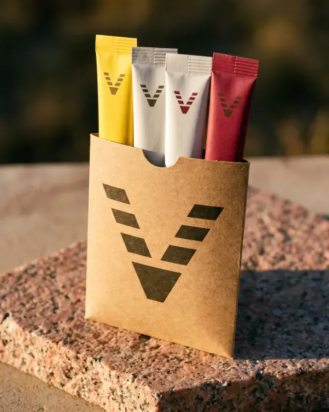 A kraft paper sleeve printed with the VitaminHill mark, with four stick packs, yellow, two white and red, standing in its open top on pink granite.